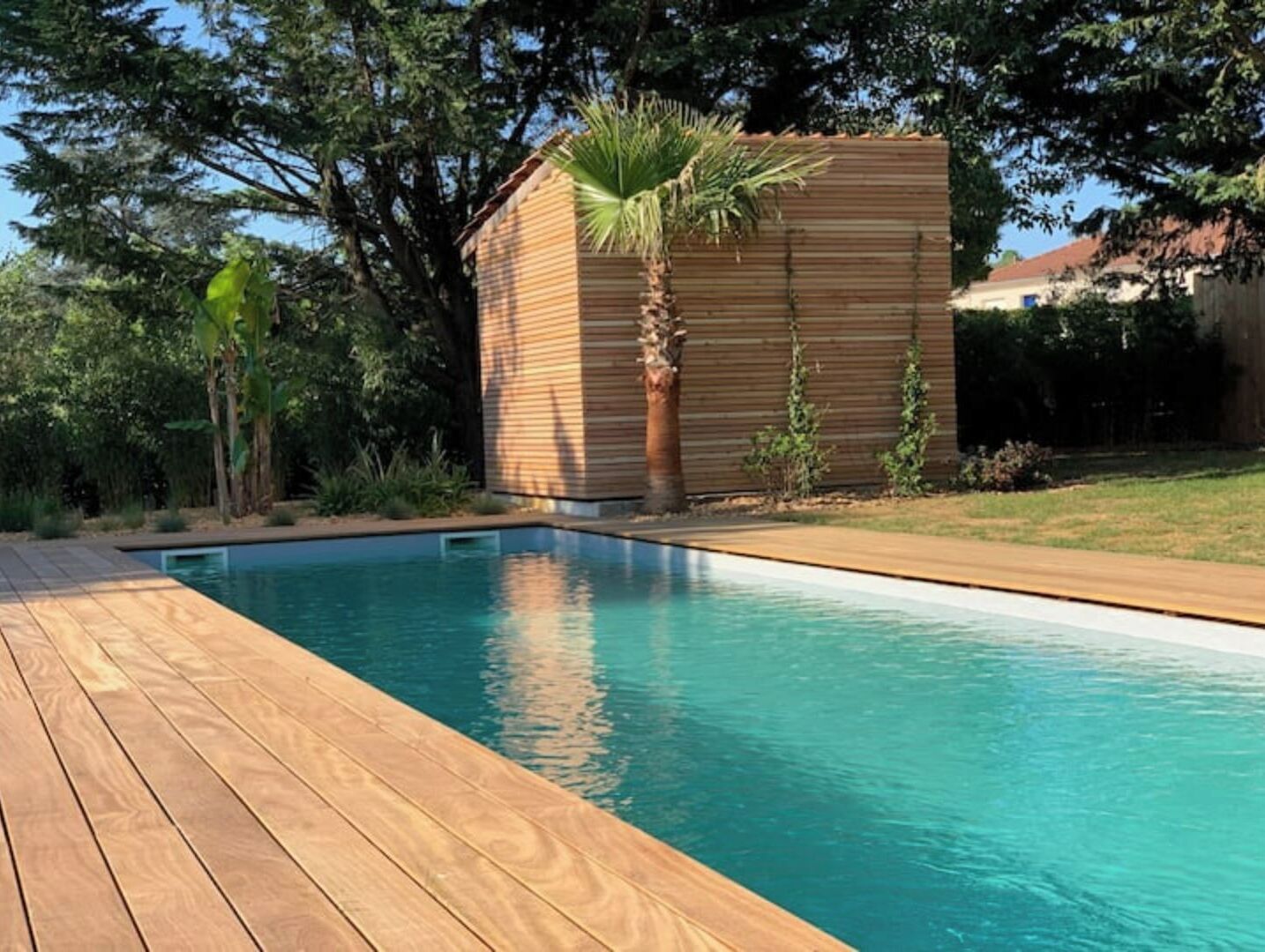 A swimming pool with a wooden deck, surrounded by greenery and a small wooden structure.