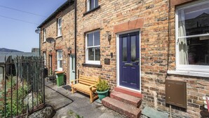 A brick house with a blue door, a bench, a potted plant, and a green trash bin. - Isabel Cottage -  a cottage that sleeps 2 guests  in 1 bedroom (Central Keswick)