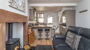 A modern kitchen with a central island, a fireplace, and a cozy seating area with a leather sofa and bar stools. - Isabel Cottage -  a cottage that sleeps 2 guests  in 1 bedroom (Central Keswick)