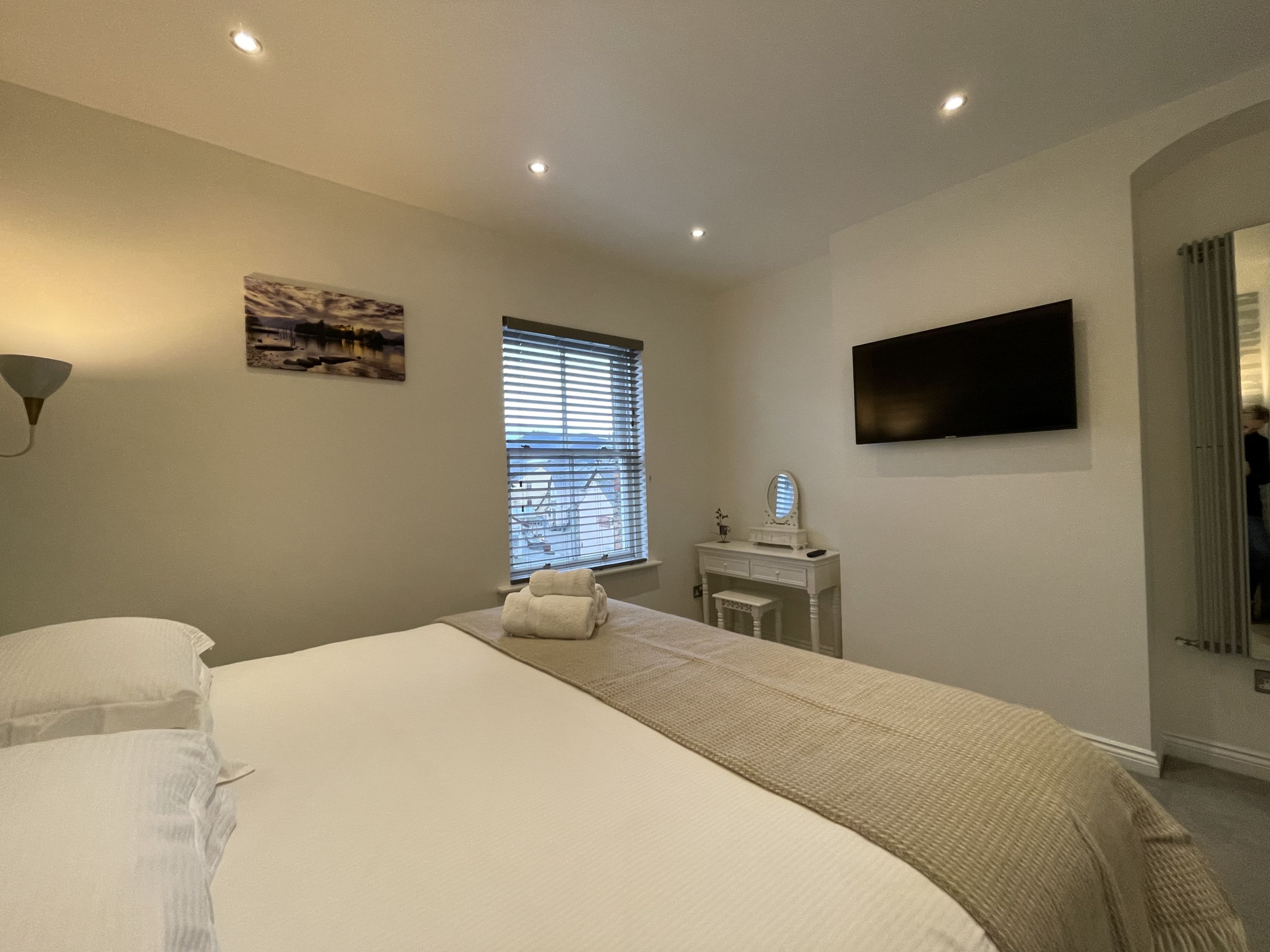 A bedroom with a bed, a vanity with a mirror, a television on the wall, and a window with blinds.