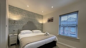 A bedroom with a bed, a nightstand, a wall-mounted lamp, a window with blinds, and a painting on the wall. - Isabel Cottage -  a cottage that sleeps 2 guests  in 1 bedroom (Central Keswick)