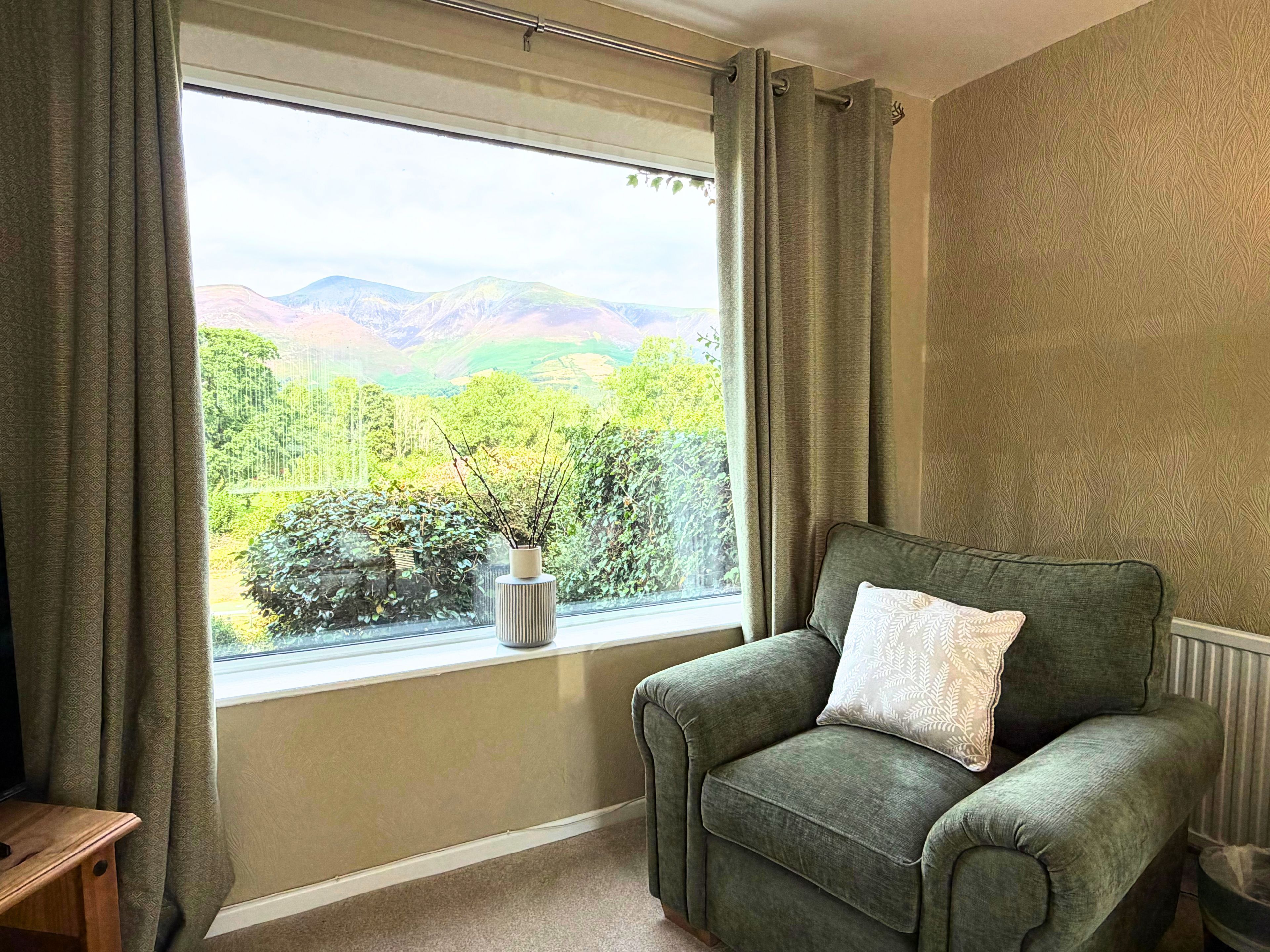 A room with a view of mountains through a window, a green armchair with a pillow, and a small table with a vase.