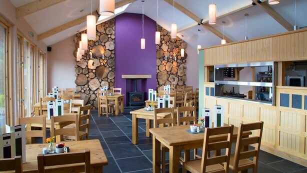 A restaurant with wooden tables and chairs, a stone wall with logs, and a kitchen area.