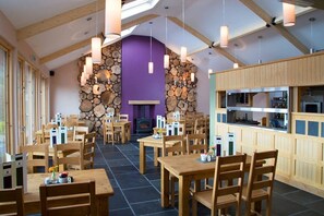 Un restaurante con mesas y sillas de madera, una pared de piedra con leñeras y un área de cocina.