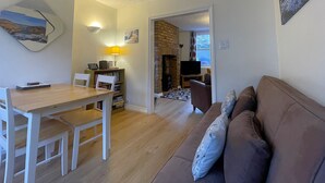 A living room with a wooden dining table, white chairs, a brown sofa, and a fireplace. - Byways -  a cottage that sleeps 4 guests  in 2 bedrooms (Central Keswick)