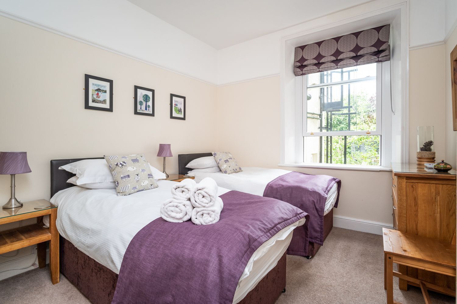 A double room with two beds, each with a purple blanket and white bedding. There are framed pictures on the wall, a window with a patterned valance, and a wooden nightstand with a lamp.
