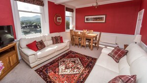 Una sala de estar con un sofá, una mesa de comedor y sillas. Hay un televisor sobre un mueble bajo y una ventana con vistas al exterior.
