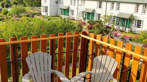 Una terraza de madera con dos sillas Adirondack, con vista a un jardín y un edificio blanco al fondo.