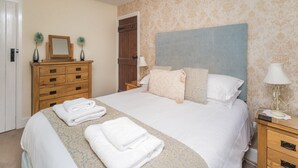 A neatly made bed with white linens, a wooden dresser with drawers, a nightstand with a lamp, and a door leading to another room. - Millside Cottage -  a cottage that sleeps 4 guests  in 2 bedrooms (Braithwaite)