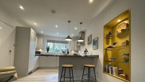 A modern kitchen with white cabinets, a central island with stools, pendant lighting, and a yellow decorative shelf with books and ceramics.