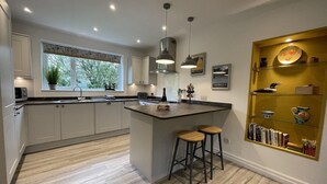 A modern kitchen with a central island, bar stools, pendant lighting, and a bookshelf with decorative items.