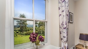 Een kamer met een groot raam met uitzicht op een groen landschap, een vaas met bloemen op een vensterbank en een lampje op een nachtkastje.