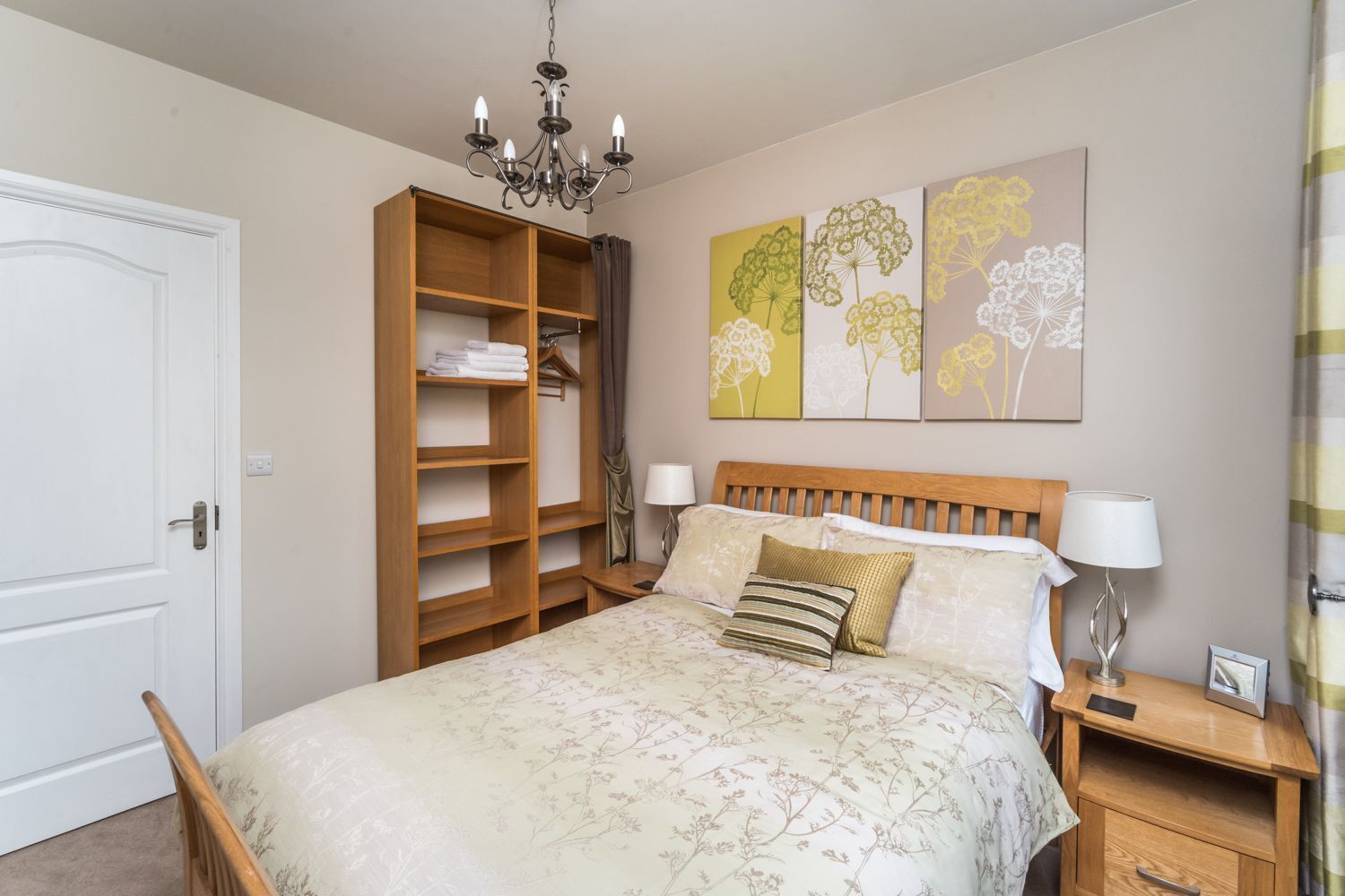 A bedroom with a bed, bedside tables, a wardrobe, a chandelier, and floral artwork on the wall.