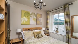 Een slaapkamer met een houten bed, bloemenbedrukte lakens, een nachtkastje met een lamp, een raam met gordijnen en uitzicht op het omliggende landschap.
