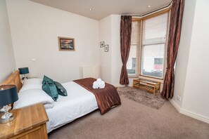 A hotel room with a bed, bedside table, lamp, and a view of the outside through a window with curtains.