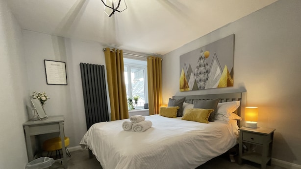 Une chambre d’hôtel bien rangée, avec un grand lit, une table de chevet, une commode, un tableau encadré au mur et une fenêtre avec des rideaux.