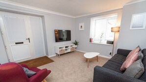 Un salon avec un canapé gris, un fauteuil rouge, un téléviseur posé sur un meuble blanc et une fenêtre avec des stores.