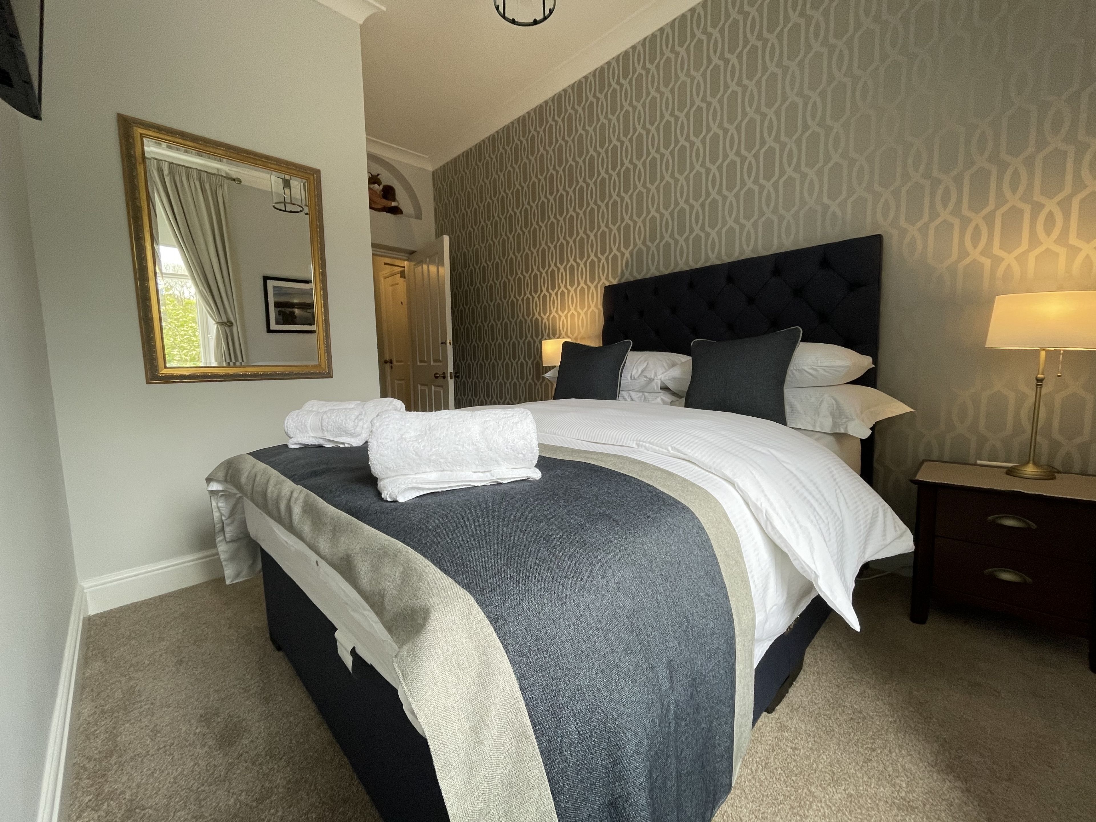 A neatly made bed with white linens and a grey and white striped blanket, a bedside table with a lamp, and a patterned wall.