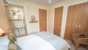 A bedroom with a bed, a wicker chair, and a bathroom with a sink and mirror. - 11 Howrah's Court -  an apartment that sleeps 4 guests  in 2 bedrooms (Central Keswick)