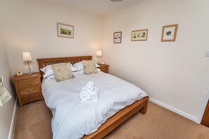 Un lit bien fait, avec du linge de lit blanc et des serviettes pliées, une tête de lit en bois, deux tables de chevet avec des lampes et trois tableaux encadrés accrochés au mur.
