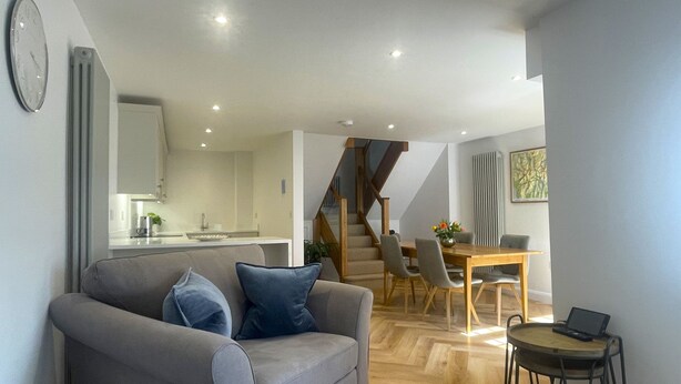 A modern living room with a grey sofa, wooden dining table, and a laptop on a small table. - Daisy Cottage -  a cottage that sleeps 4 guests  in 2 bedrooms (Central Keswick)