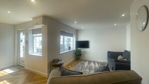 A spacious living room with a large sofa, a flat-screen TV mounted on the wall, and a window providing natural light. - Daisy Cottage -  a cottage that sleeps 4 guests  in 2 bedrooms (Central Keswick)