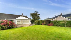 Een woonhuis met een verzorgd gazon, een tuinkamer en diverse struiken en bloeiende planten.