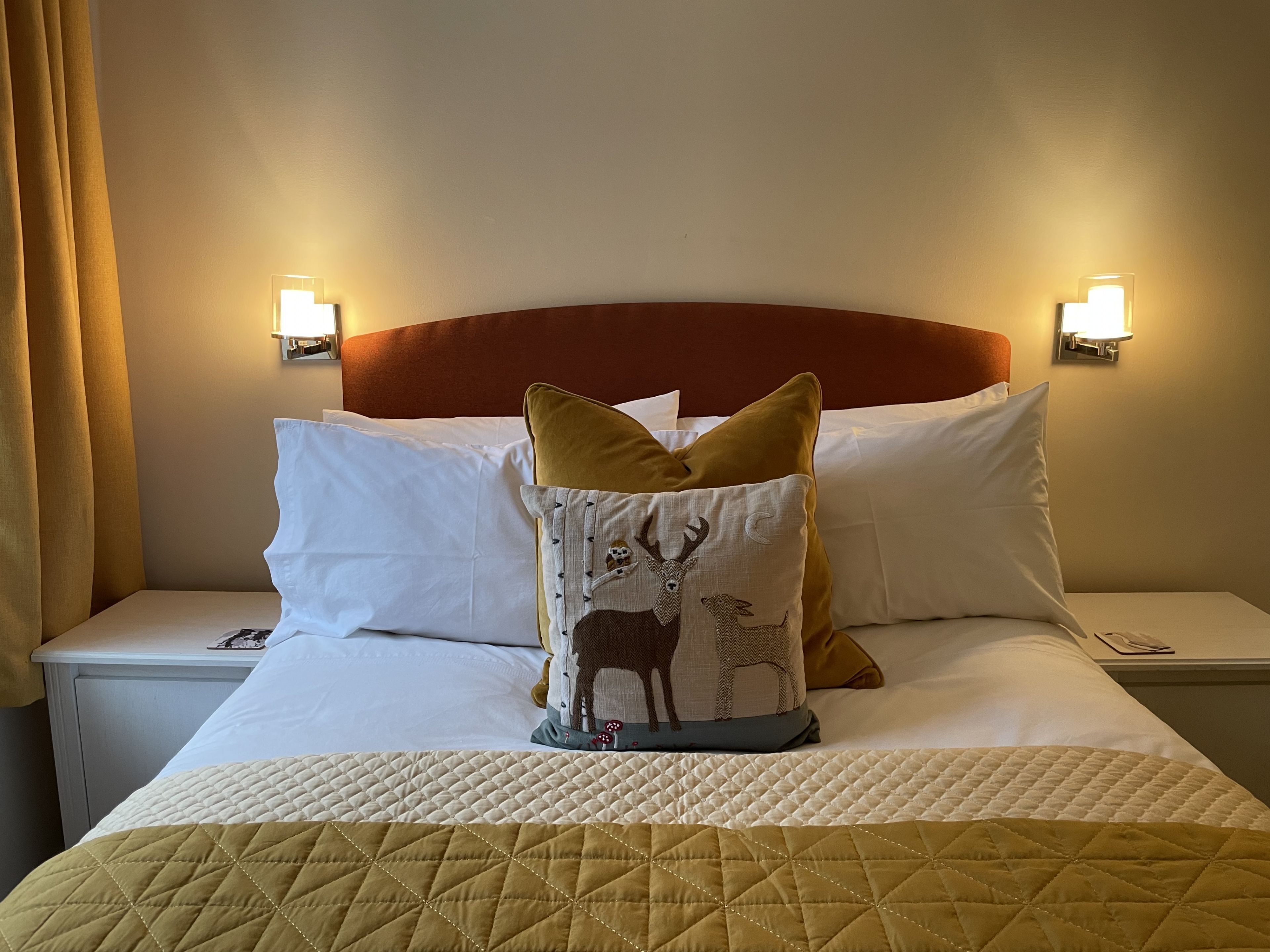 Habitación de hotel con cama, ropa blanca, cabecero acolchado y almohadones decorativos, incluyendo uno con diseño de ciervo.