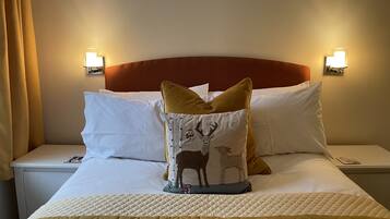 Habitación de hotel con cama, ropa blanca, cabecero acolchado y almohadones decorativos, incluyendo uno con diseño de ciervo.