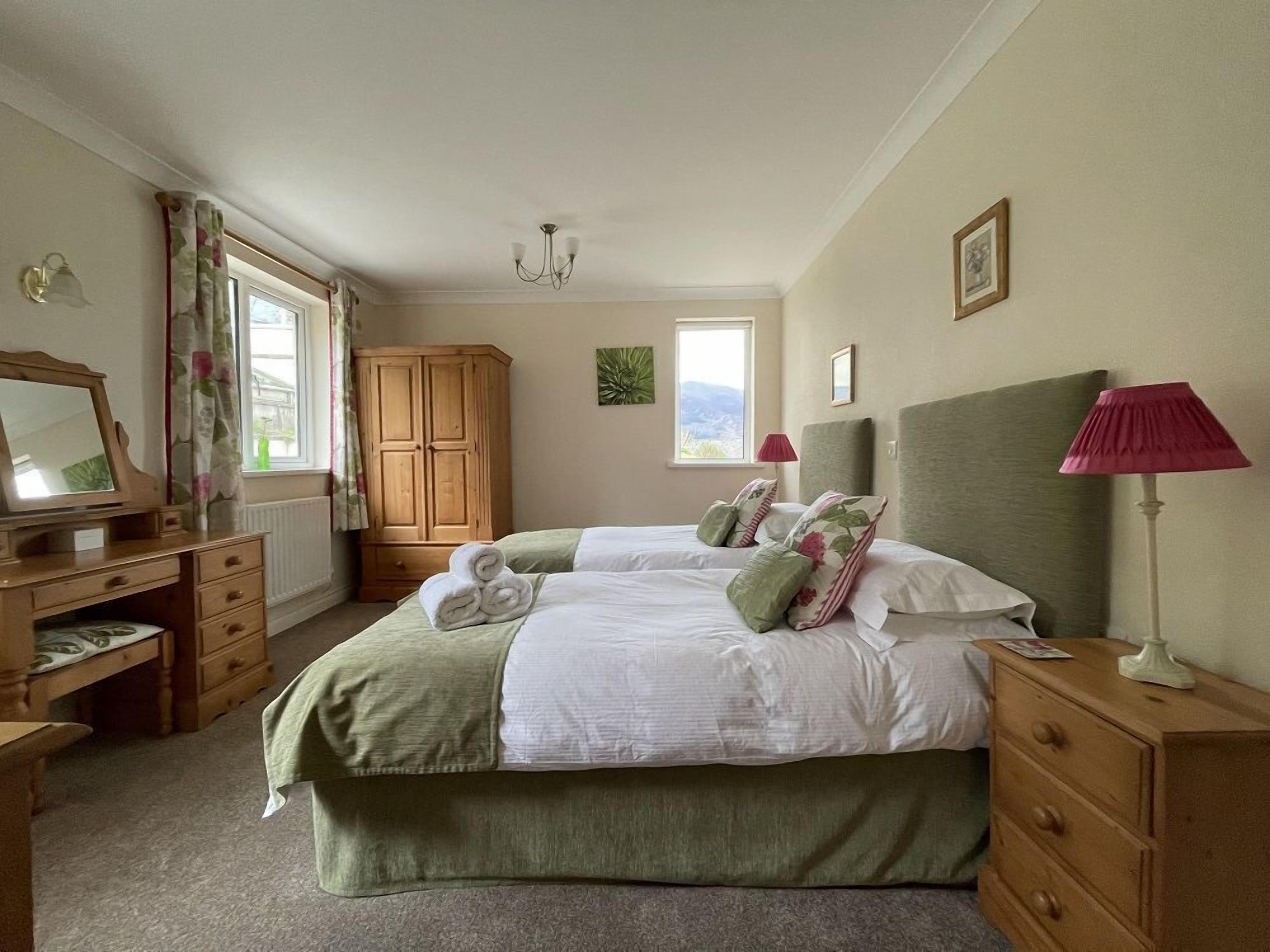 A bedroom with two beds, a dresser, a vanity with a mirror, a window with curtains, a wooden wardrobe, and a lamp.