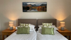 Een tweepersoonsbed met witte lakens en groene kussens, met aan weerszijden nachtkastjes met lampen, tegen een muur met een lijstwerklandschap.