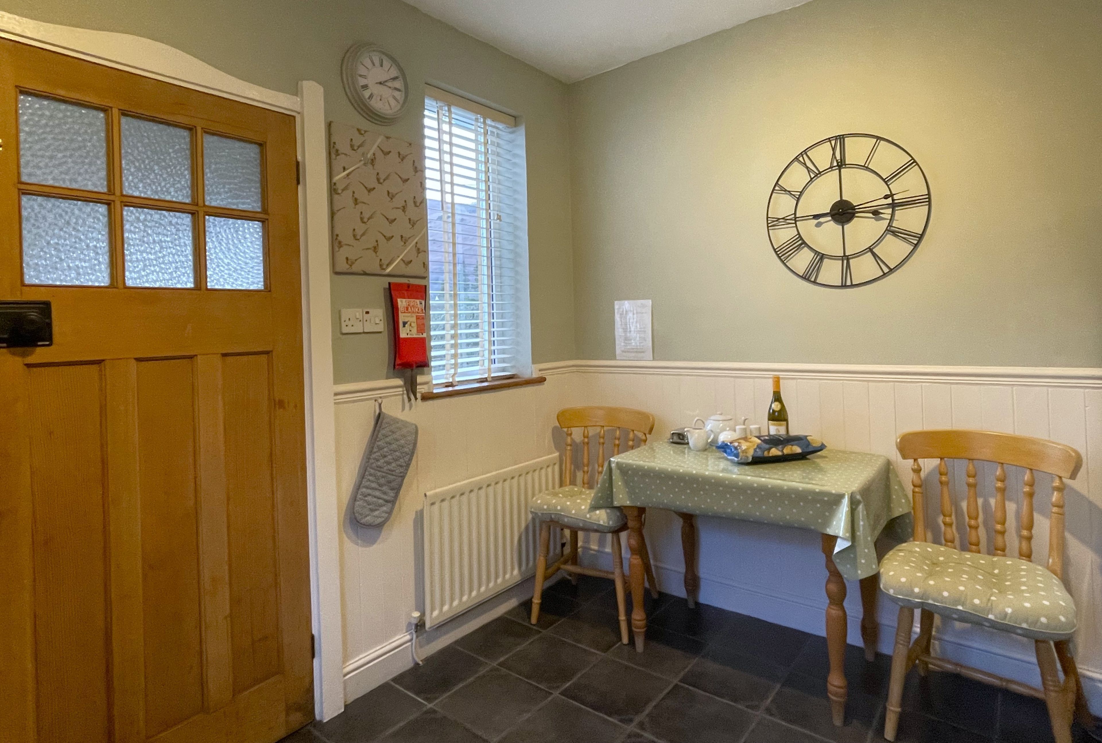 A dining area with a table, chairs, a clock, and a window with blinds.