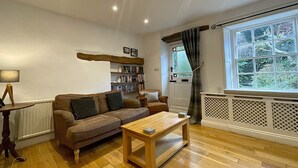 A cozy living room with a wooden floor, a brown sofa, a coffee table, a bookshelf, and a window with curtains. - Barf Cottage -  a cottage that sleeps 5 guests  in 3 bedrooms (Portinscale)