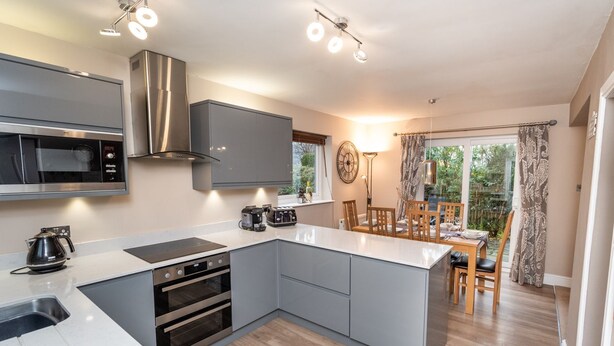 A modern kitchen with grey cabinets, a built-in oven, a microwave, and a coffee maker. There is a dining area with a table and chairs, and a large window with curtains. - Appletree -  a house that sleeps 6 guests  in 3 bedrooms (Portinscale)