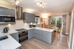 Eine moderne Küche mit grauen Schränken, einem eingebauten Backofen, einer Mikrowelle und einer Kaffeemaschine. Es gibt einen Essbereich mit Tisch und Stühlen sowie ein großes Fenster mit Vorhängen.