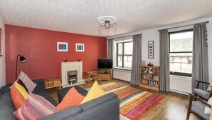 A living room with a fireplace, sofa, TV, bookshelf, and large windows. - Alban Rise -  a maisonette that sleeps 5 guests  in 3 bedrooms (Central Keswick)