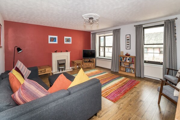 A living room with a fireplace, sofa, TV, bookshelf, and large windows. - Alban Rise -  a maisonette that sleeps 5 guests  in 3 bedrooms (Central Keswick)