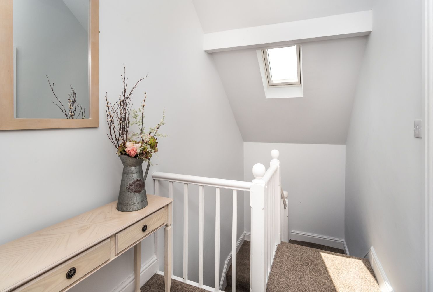 Une chambre moderne et bien éclairée, située sous les combles, avec un puits de lumière, une commode en bois et un vase rempli de fleurs.