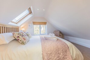 A bedroom with a sloped ceiling, a skylight, a bed with floral pillows, a bedside table with towels, and a window with blinds.