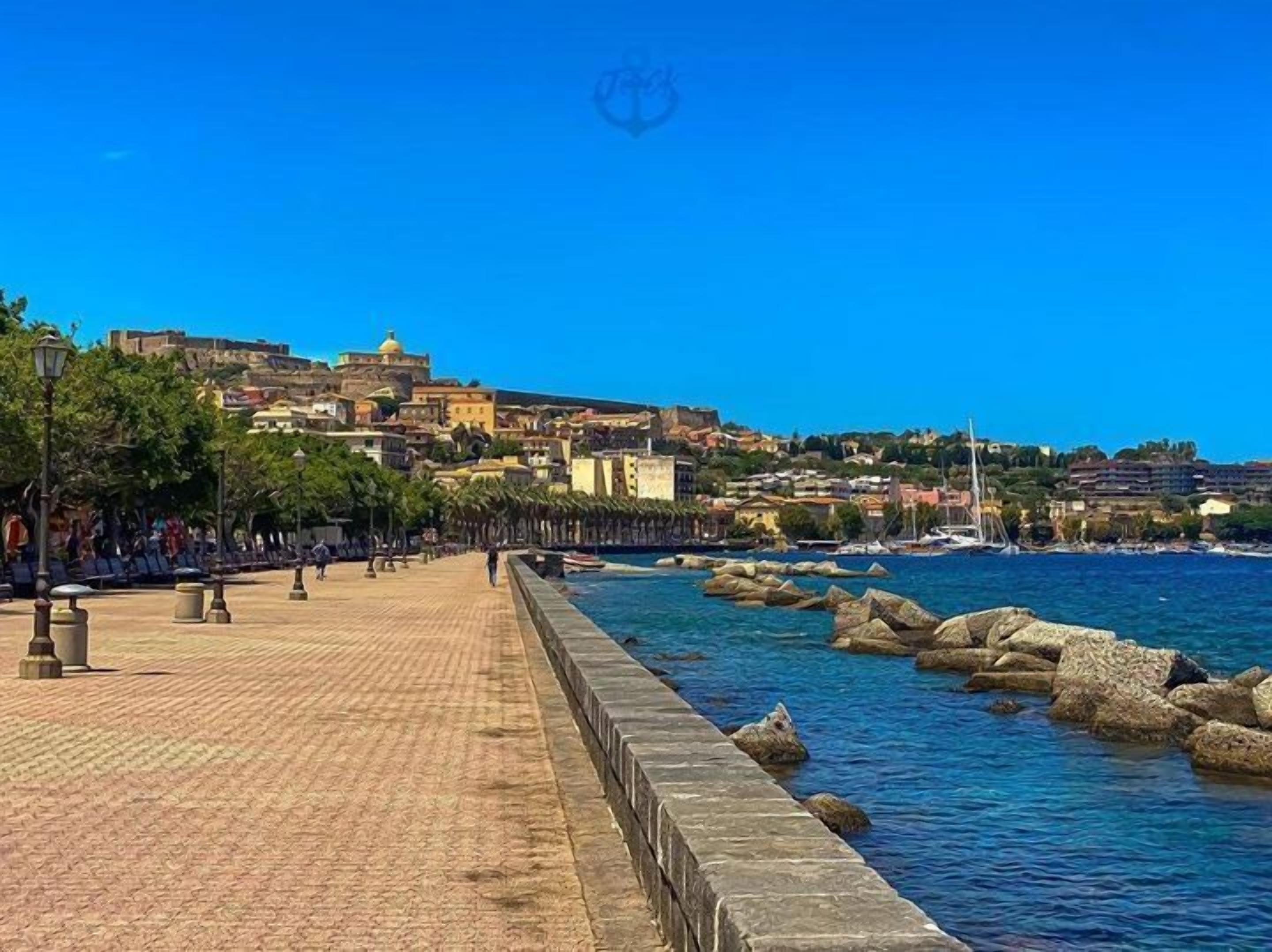 Um calçadão costeiro com um pavimento de pedra, vista para o mar e edifícios ao fundo.