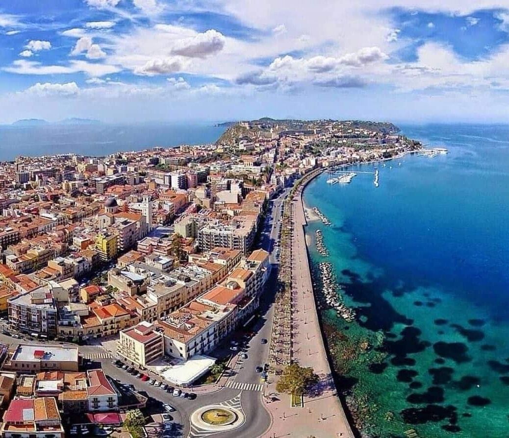 Vista aerea di una città costiera con un importante lungomare, una rotonda centrale e un mix di edifici residenziali e commerciali.