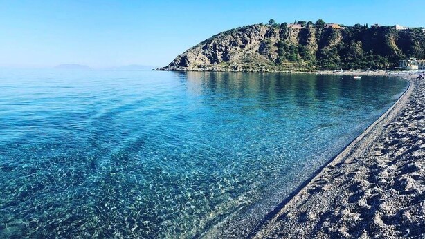 A clear view of a beach with a rocky cliff and a clear blue sea.