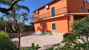 A two-story building with a red facade, a balcony with green shutters, and a paved courtyard.