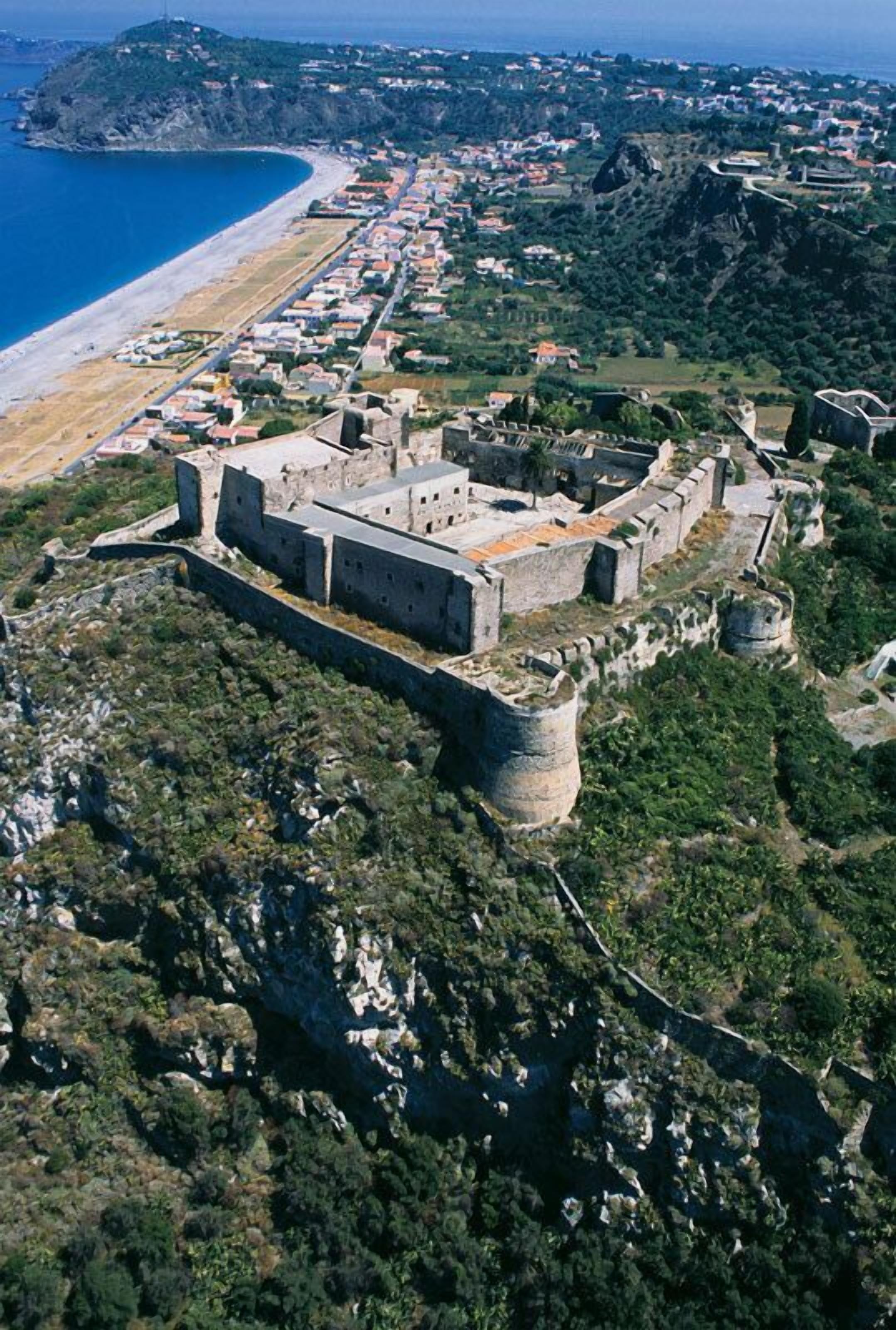 Vista aérea de uma vila costeira com uma importante fortificação histórica numa colina, praia e zonas residenciais.