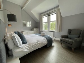 Ein modernes Schlafzimmer mit einem großen Bett, einem Nachttisch, einem Sessel, einem Fenster mit Vorhängen und einem Spiegel.