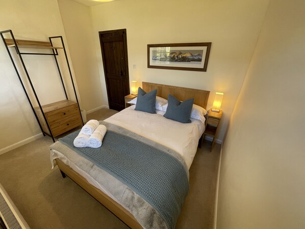 Een net opgemaakt bed met een grijze deken en kussens, een houten hoofdbord, een ingelijst beeld aan de muur en een houten nachtkastje met een lamp.