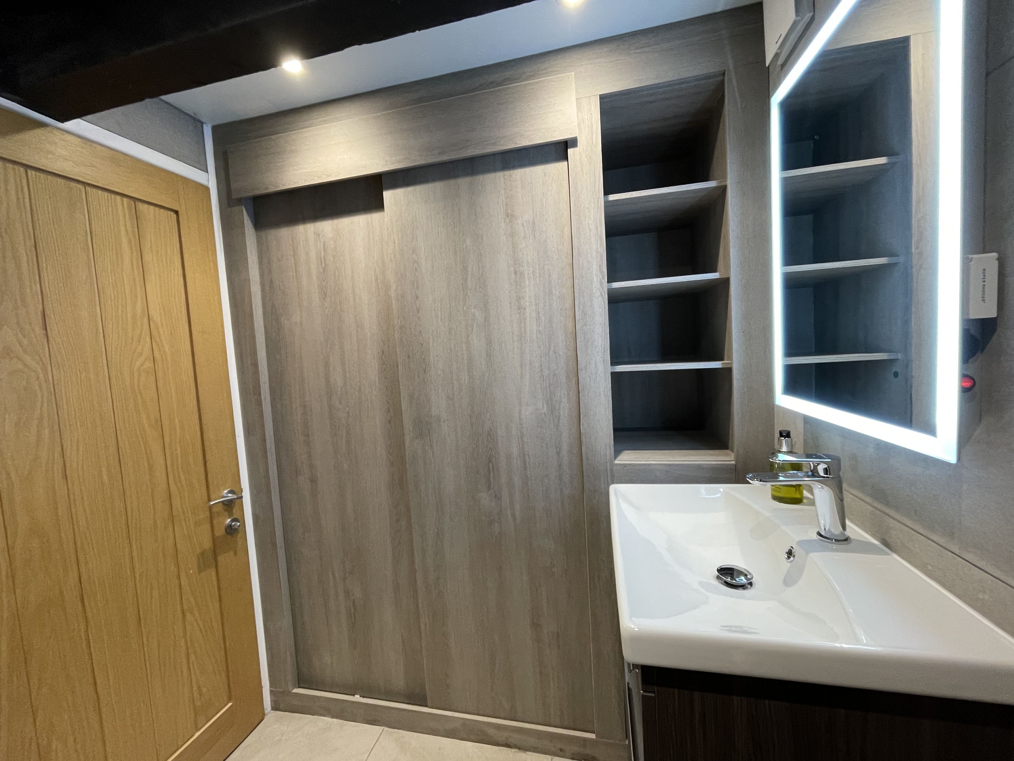 Een badkamer met een houten deur, een witte wastafel en een spiegel met ingebouwde verlichting.