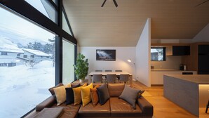 Ein modernes Wohnzimmer mit großem Fenster, einer Couch mit Kissen, einem Essbereich mit Stühlen und einer Küchenzeile.