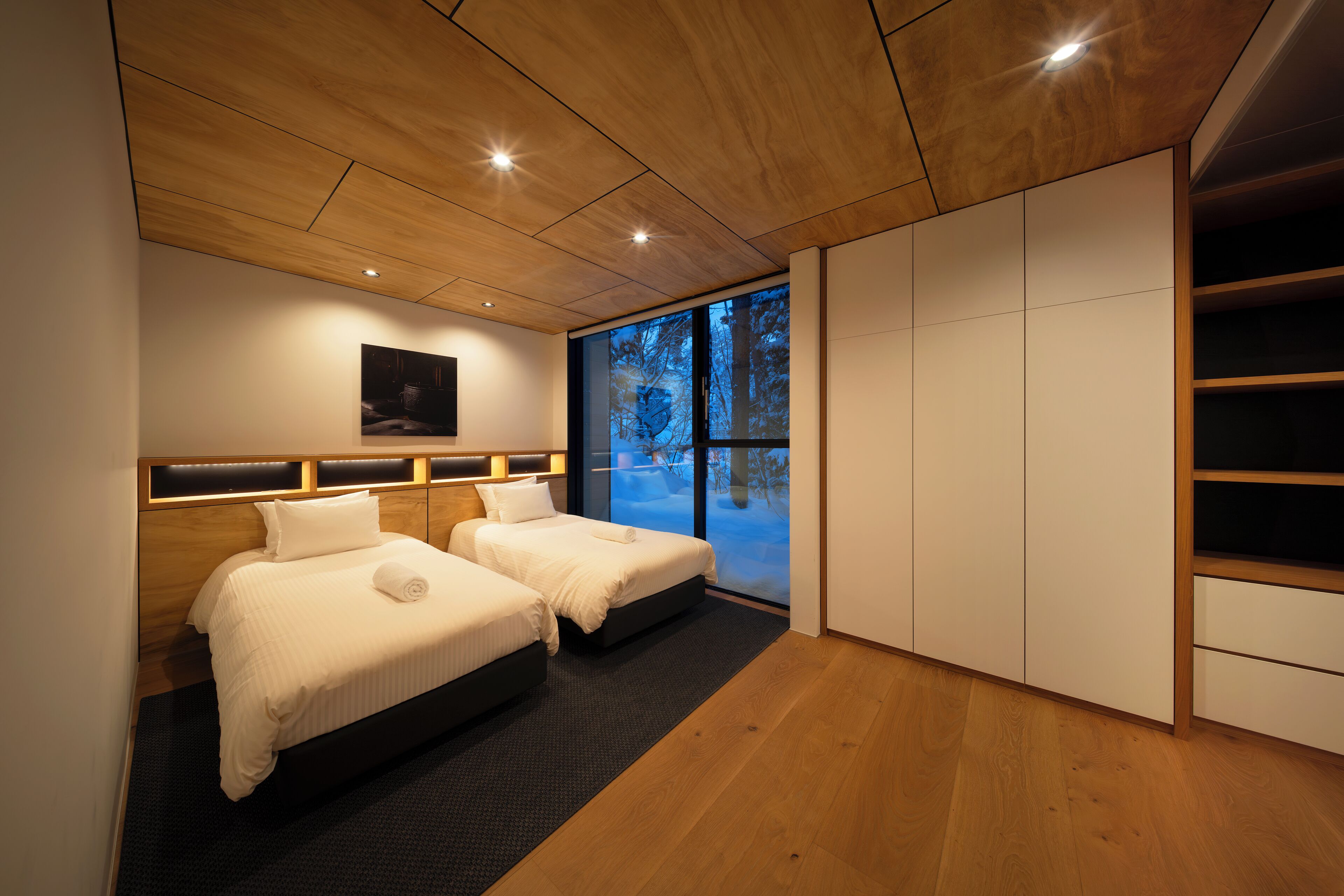A modern hotel room with two beds, a large window with a view of trees, and a built-in wardrobe.