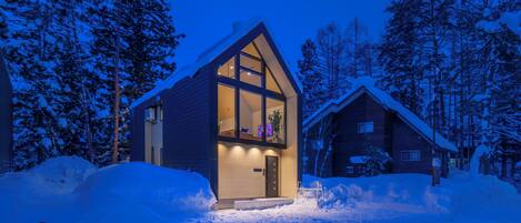 A modern house with a large glass window and a door, surrounded by snow and trees.